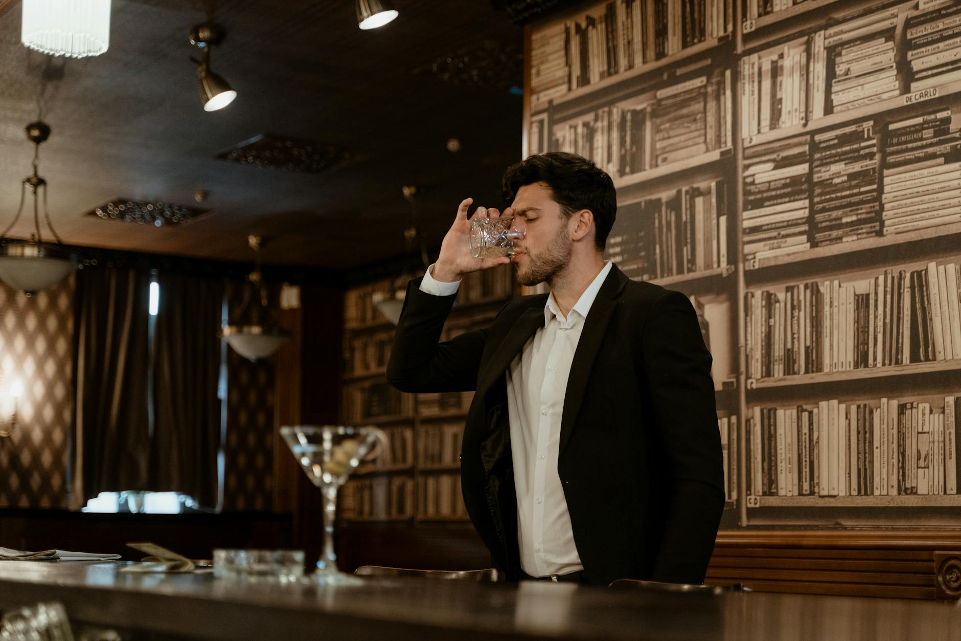 Elegancki mężczyzna pije drinka samotnie w stylowym barze z książkami Elegancki mężczyzna pije drinka samotnie w stylowym barze z książkami